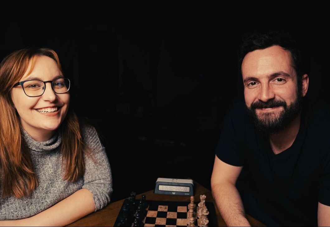 A woman in a grey shirt and glasses and a man with a beard in a black shirt smile at the camera. They are seated on opposite sides of a chessboard with a clock, ready to play a game.